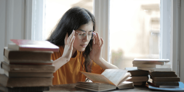 A young woman wearing glasses surrounded by books. She has her hands to her temples in a sign of stress.