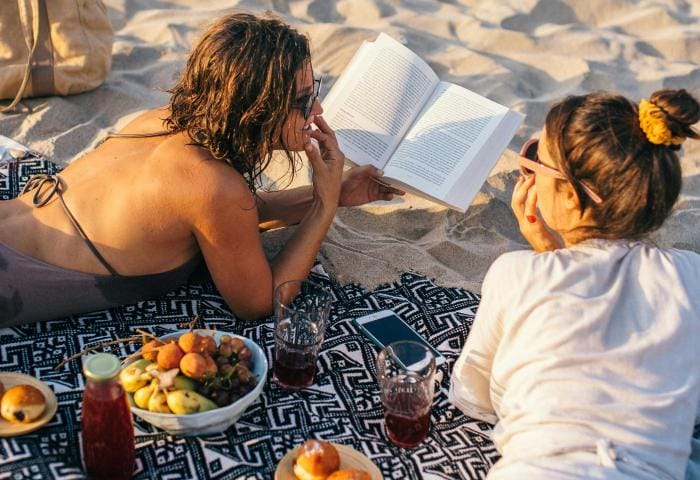 Two women reading a book on a beach with snacks